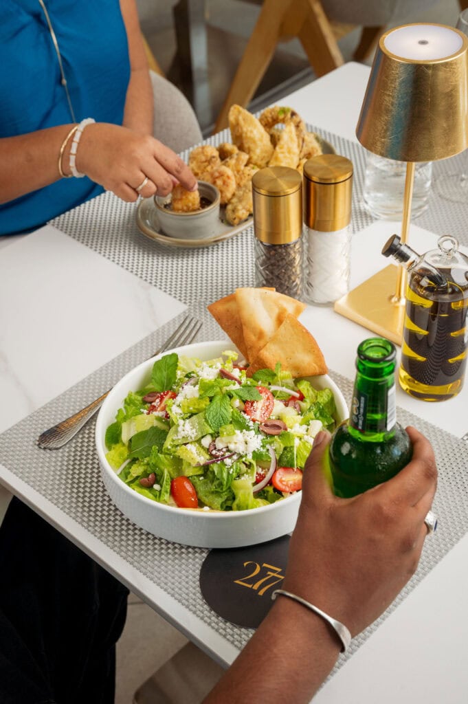 "Fresh Mediterranean Salad with romaine lettuce, feta cheese, olives, and tomatoes, served in a white bowl with pita chips and a bottled beverage at 277 Restaurant in Trinidad.