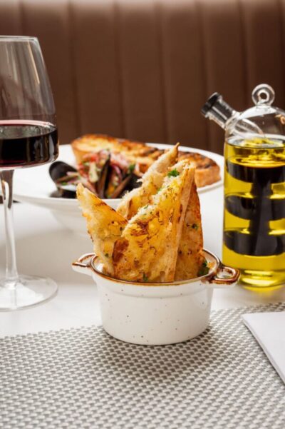 Close-up of crispy, herbed bread or garlic toast served in a small pot, with a red wine glass and olive oil on the side, highlighting an appetizer or side dish at 277 Restaurant.