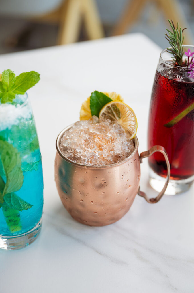 Variety of 277 Restaurant signature cocktails: a copper mug with an Infused Vodka, Bois Bande-inspired drink, beside a Red Wine Sangria and a blue Coconut-Pineapple-Mint Mojito, showcasing the diverse bar menu