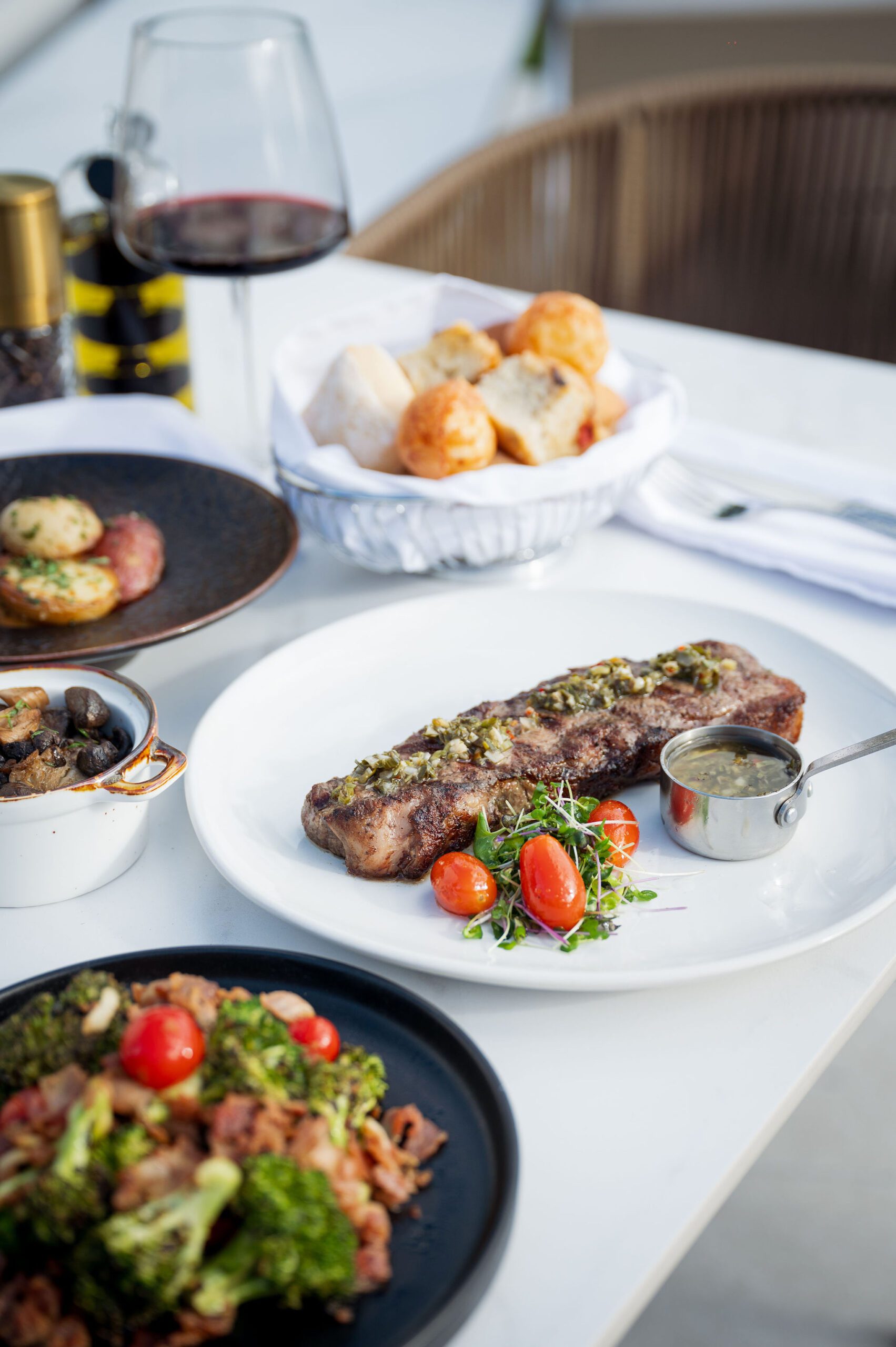 Outdoor dining table featuring a grilled NY Striploin steak, red wine, bread basket, potatoes, and broccoli side dishes, showcasing the complete meal experience at 277 Restaurant